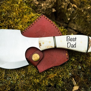 May include: A small, personalized axe with a white handle and a leather sheath. The handle is engraved with the words "Best Dad".