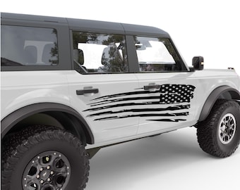 Könnte beinhalten: Ein weißer Ford Bronco mit einem schwarzen und weißen, abgenutzten Aufkleber mit der amerikanischen Flagge an der Seite. Der Aufkleber befindet sich an der Fahrertür und erstreckt sich von der Vordertür bis zur Hintertür.