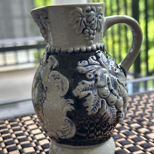 May include: A gray and blue ceramic pitcher with a handle. The pitcher features embossed grape and leaf designs, along with other decorative relief patterns. The base is a lighter shade of gray, and the pitcher has a small spout.