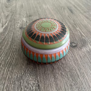 Puede incluir: Pequeña caja decorativa redonda con tapa. La tapa presenta un diseño de sol en verde, naranja y negro. El cuerpo de la caja es turquesa y naranja con un patrón geométrico. La caja tiene aproximadamente 7,6 cm de diámetro.