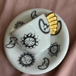 Puede incluir: Un juego de taza de espresso y platillo de cerámica blanca con ilustraciones de líneas negras de soles y lunas crecientes. La taza tiene un asa dorada distintiva con forma de abanico o ala, añadiendo un toque metálico al diseño.