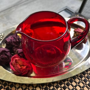 May include: A ruby red glass creamer with a rounded body and a curved handle sits on a silver tray. Dried red roses are arranged around the creamer. The creamer is approximately 10 cm tall.