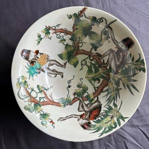 May include: A cream-colored ceramic plate with a detailed illustration of monkeys in a tree. The monkeys are depicted with hats and holding objects. The tree branches and leaves are in shades of brown, green, and yellow. The plate has a slightly raised edge.