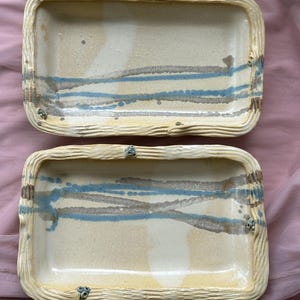 Puede incluir: Dos bandejas rectangulares de cerámica con base color crema, que presentan bordes texturizados similares a la madera en un tono amarillo pálido. Las superficies interiores están decoradas con pinceladas horizontales y diagonales en azul y marrón.