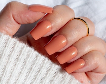 Op de afbeelding: Close-up van een hand met lange, vierkante, perzik-kleurige kunstnagels. Een dunne gouden ring siert de ringvinger. De hand rust op een witte, geribbelde gebreide trui. De nagels hebben een glanzende afwerking.