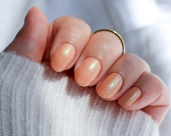 Op de afbeelding: Close-up van een hand met perzikkleurige nagellak met een glinsterende afwerking. De nagels zijn gemanicuurd en de lak is gelijkmatig aangebracht.