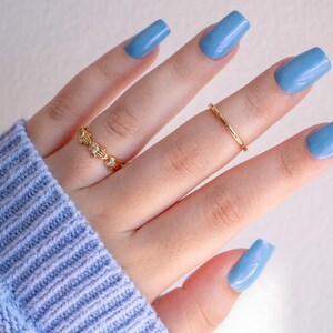 Op de afbeelding: Een hand met lichtblauwe nagellak op elke vinger. De hand draagt twee gouden ringen.