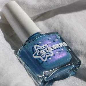 Op de afbeelding: Een klein flesje blauwe nagellak met een witte ster en het woord "STEORRA" op het etiket. Het flesje staat op een wit oppervlak.