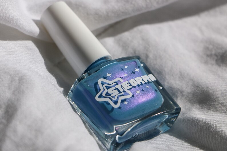 Op de afbeelding: Een klein flesje blauwe nagellak met een witte ster en het woord "STEORRA" op het etiket. Het flesje staat op een wit oppervlak.