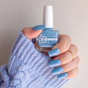 Op de afbeelding: Een hand met een lichtblauwe kabelgebreide trui houdt een flesje lichtblauwe nagellak vast met een witte ster en het woord "STEORRA" op het etiket.