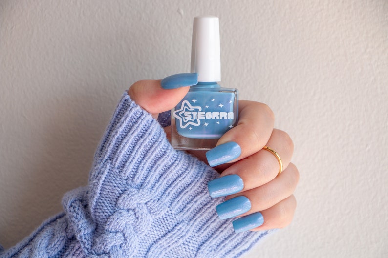 Op de afbeelding: Een hand met een lichtblauwe kabelgebreide trui houdt een flesje lichtblauwe nagellak vast met een witte ster en het woord "STEORRA" op het etiket.
