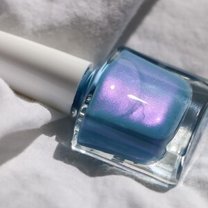 Op de afbeelding: Een flesje lichtblauwe nagellak met een glinsterende afwerking. De fles is van helder glas en heeft een witte dop.