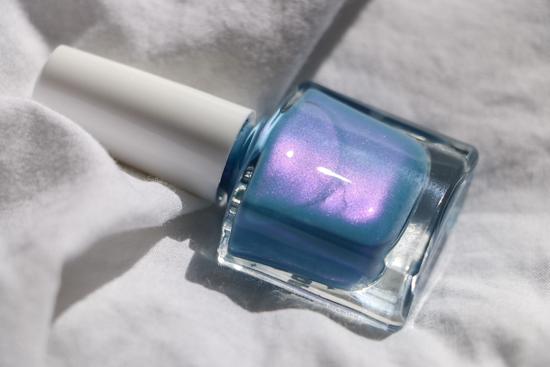 Op de afbeelding: Een flesje lichtblauwe nagellak met een glinsterende afwerking. De fles is van helder glas en heeft een witte dop.