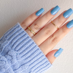 Op de afbeelding: Een hand met lichtblauwe nagellak op elke vinger. De hand draagt twee gouden ringen, een met kleine heldere steentjes en een eenvoudige band.