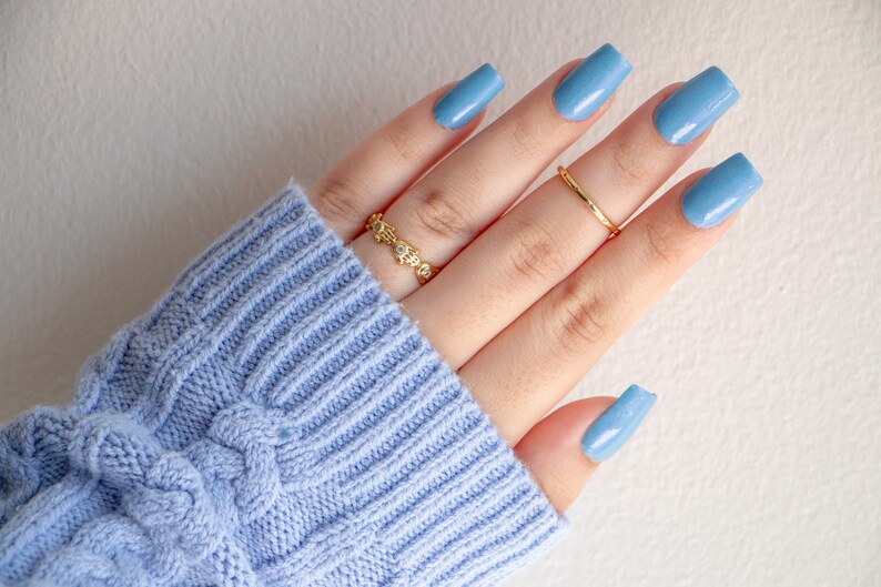Op de afbeelding: Een hand met lichtblauwe nagellak op elke vinger. De hand draagt twee gouden ringen, een met kleine heldere steentjes en een eenvoudige band.