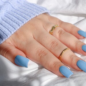 Op de afbeelding: Een hand met lichtblauwe vierkante nagellak en twee gouden ringen. De hand rust op een wit oppervlak.