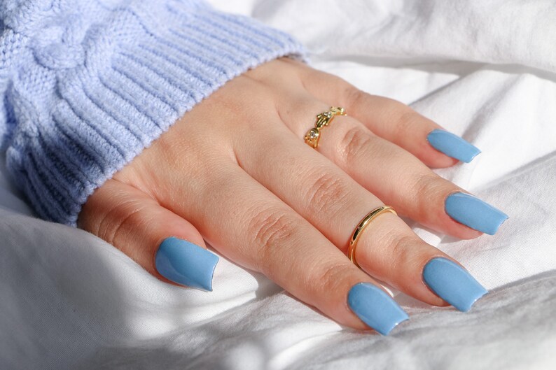 Op de afbeelding: Een hand met lichtblauwe vierkante nagellak en twee gouden ringen. De hand rust op een wit oppervlak.