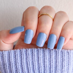 Op de afbeelding: Een hand met lichtblauwe nagellak op elke vinger. De nagels zijn kort en vierkant gevormd. De hand draagt een gouden ring aan de ringvinger.