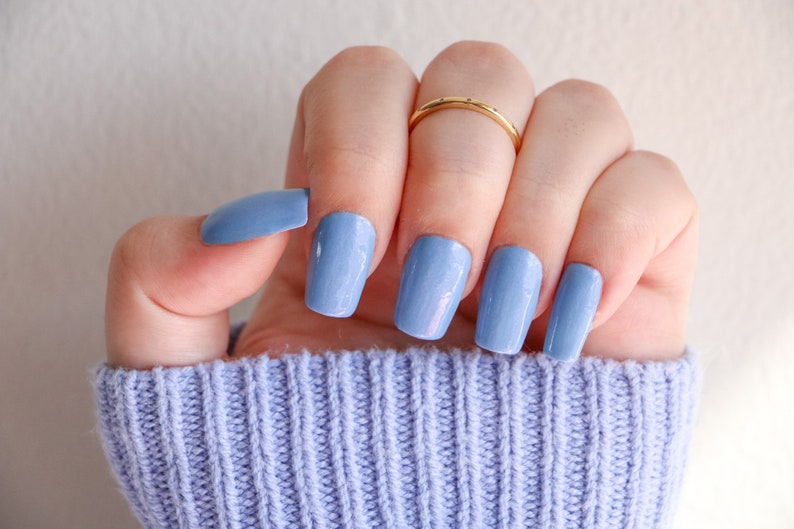 Op de afbeelding: Een hand met lichtblauwe nagellak op elke vinger. De nagels zijn kort en vierkant gevormd. De hand draagt een gouden ring aan de ringvinger.