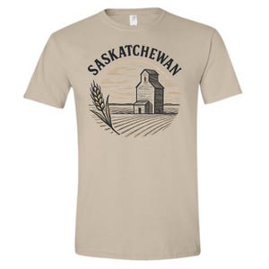 May include: Beige t-shirt with a graphic of a grain elevator and wheat, with the word "Saskatchewan" in a circular design. The design is in black and beige, with a vintage aesthetic.
