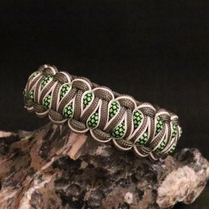 May include: A handmade paracord bracelet in shades of gray, white, and neon green. The bracelet features a woven design with a series of interconnected knots. The accessory is displayed on a textured rock surface against a dark background.
