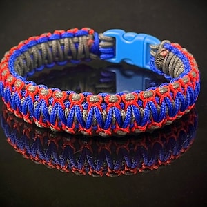 May include: A blue, red, and gray braided bracelet with a blue plastic clasp. The bracelet is made of paracord and has a woven design.