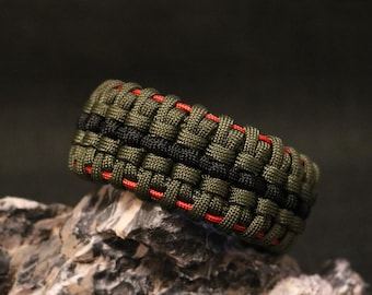 Könnte beinhalten: Ein grünes und schwarzes Paracord-Armband mit roten Akzenten. Das Armband ist in einem komplexen Muster gewebt.