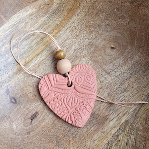 May include: A terracotta heart-shaped ornament with a detailed patterned design, hanging from a twine cord with wooden beads.