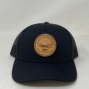 Op de afbeelding: Zwarte truckerpet met een mesh achterkant en een gebogen klep. De pet heeft een cirkelvormige patch met de woorden "Camp Crystal Lake Camp Counselor" rond een berg- en meer-afbeelding.