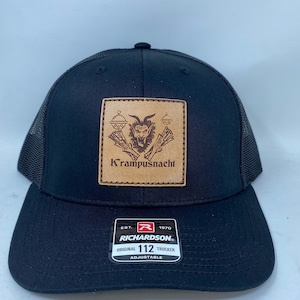 Op de afbeelding: Zwarte truckercap met een bruine leren patch met een Krampusnacht-ontwerp. De patch heeft een gestikte rand en de tekst "Krampusnacht". De cap is verstelbaar en heeft een mesh-rug.
