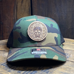 May include: A camouflage trucker hat with a brown leather patch featuring a Japanese demon mask design. The patch has a wave pattern around the mask. The hat has a black mesh back and a black adjustable strap with a black label that reads "Richardson 112 Original Trucker Adjustable".