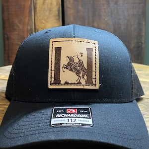 Op de afbeelding: Zwarte truckerpet met een mesh achterkant en een vierkante leren patch. De patch toont een bruine afbeelding van een persoon op een paard tussen twee pilaren. De pet heeft het Richardson 112 Trucker label.