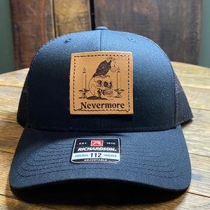 May include: Black trucker hat with a brown leather patch featuring a skull with a raven perched on top and the text "Nevermore". The hat has a black mesh back and a Richardson 112 label.