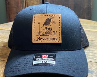May include: Black trucker hat with a brown leather patch featuring a skull with a raven perched on top and the text "Nevermore". The hat has a black mesh back and a Richardson 112 label.