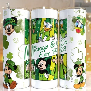 Könnte beinhalten: Drei weiße Edelstahlbecher mit grünen, gelben und schwarzen Comicbildern von Mickey Mouse, Minnie Mouse, Donald Duck, Goofy und Pluto, die grüne Kleeblatt-Hüte tragen. Die Becher haben einen weißen Hintergrund mit grünen Kleeblatt-Designs. Der mittlere Becher trägt den Text "Mickey & Co Est 1928".