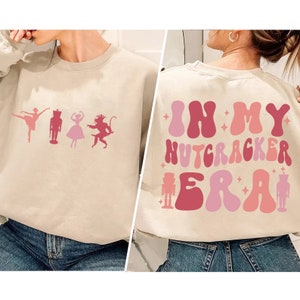 Puede incluir: Una sudadera beige con un diseño gráfico rosa y rojo en la parte delantera y trasera. El diseño delantero presenta una bailarina, un cascanueces, una princesa y un mono. El diseño trasero presenta el texto "In My Nutcracker Era" en una fuente retro.