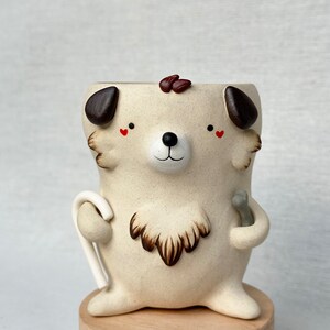 May include: A ceramic planter shaped like a dog with brown ears, a brown nose, and red hearts for eyes. The planter is holding a white candy cane in one hand and a gray stick in the other.