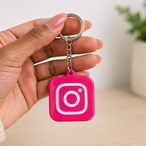 Pode incluir: Um chaveiro rosa com o logotipo branco do Instagram. Possui uma corrente e um chaveiro prateados. O chaveiro é segurado por uma mão.