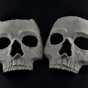 Könnte beinhalten: Zwei graue Totenkopfmasken mit Augen- und Nasenöffnungen werden vor einem schwarzen Hintergrund gezeigt. Die Masken weisen detaillierte Merkmale auf, darunter Zähne und Gesichtsriss, was auf ein Halloween- oder Kostümthema hindeutet.