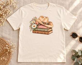 May include: A cream-colored short-sleeve t-shirt featuring a graphic print of a tan cowboy hat resting on a stack of three books, adorned with a cluster of white and pink daisies and green foliage on the left side of the stack.