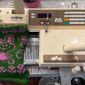 May include: A green and pink knitted fabric with a bunny design is on a Brother knitting machine. The machine is off-white with a control panel and a handle. The knitted fabric is green with pink bunny and floral patterns.