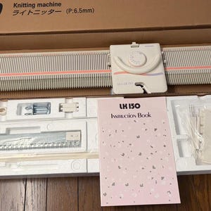 May include: A beige LK 150 knitting machine with a carriage, housed in a cardboard box with styrofoam inserts. The set includes a pink instruction book labeled LK 150, various plastic tools, metal components, and a long needle bed with pink accents.
