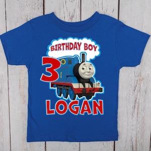 May include: A royal blue short-sleeve t-shirt featuring a graphic of a blue steam engine train. The design includes the red number 3, the text BIRTHDAY BOY in a white cloud shape, and the name LOGAN in large red letters below the train graphic.