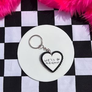 Puede incluir: Un llavero blanco en forma de corazón con las palabras "WE'LL BE ALRIGHT" en escritura negra, enmarcado por un borde negro festoneado. El llavero está sujeto a un anillo plateado y descansa sobre una base circular blanca. El fondo presenta un patrón de cuadros blancos y negros y piel sintética rosa.