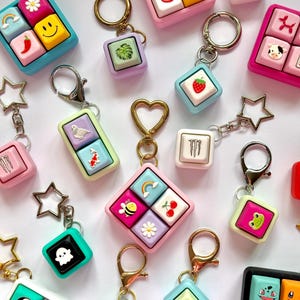 May include: A collection of colorful mechanical keyboard keycap keychains featuring various icons including rainbows, smiley faces, strawberries, animals, flowers, and logos. Each keycap is mounted on a metal ring, some with star or heart-shaped clasps.