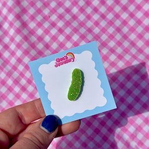 Puede incluir: Un broche verde brillante en forma de pepinillo sobre una tarjeta azul claro con el texto "Gee's Jewellery" y un logotipo de arcoíris. La tarjeta se sostiene sobre un fondo a cuadros rosa y blanco.