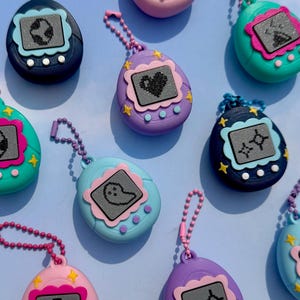 May include: A collection of colorful, retro-style handheld game keychains. Each keychain features a different color body, a small screen with a pixelated graphic, and a matching beaded chain. The keychains are in shades of pink, blue, purple, and teal.