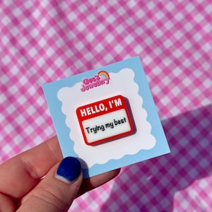 May include: A red and white enamel pin with the text "HELLO, I'M Trying my best" on a light blue card. The card has a white cloud-shaped border and the Gee's Jewellery logo. The background is a pink and white gingham pattern.