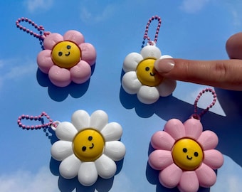 May include: Four daisy-shaped keychains in pink and white with yellow centers and smiling faces. Each keychain has a pink beaded chain. The background is a light blue sky with white clouds.