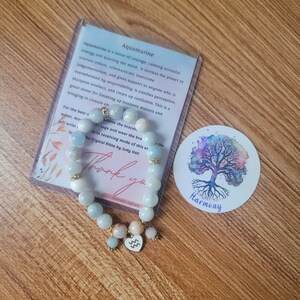 May include: Aquamarine bracelet with a heart charm featuring the astrological symbol for Aquarius. The bracelet is made with light blue, white, and pink beads. The background features a wooden surface and a sticker with a tree and the word "Harmony".